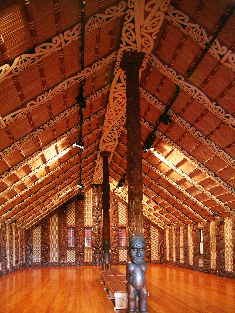 The carved interior of the wharenui Te Whare Rūnanga, with a carved centre post and woven wall panels lit from above